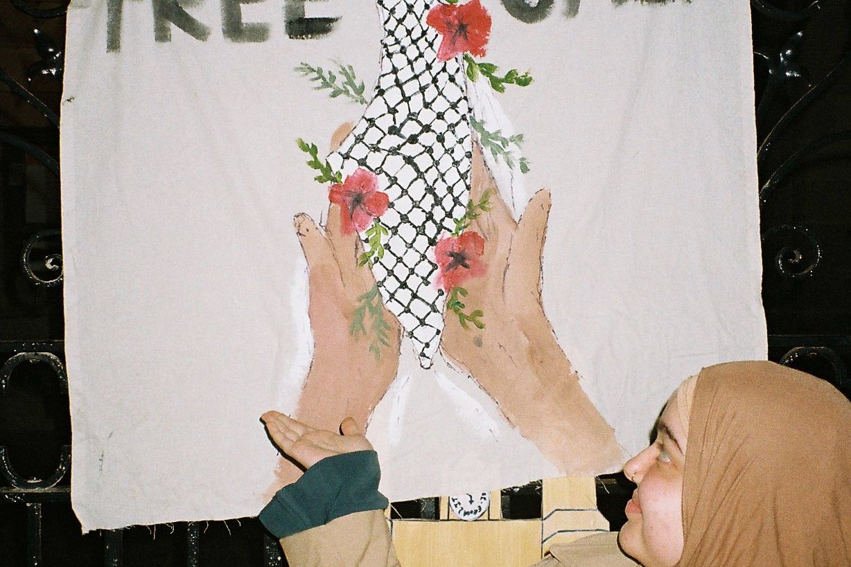 A person showing a banner which has the text "FREE GAZA" with hands uplifitng a map of Palestine covered in the kuffiyeh pattern